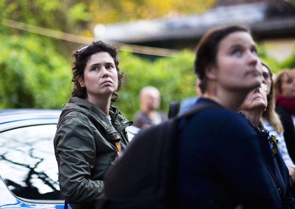 Kobieta w zielonej kurtce spogląda w g&oacute;rę. W tle rozmyte postaci innych os&oacute;b.