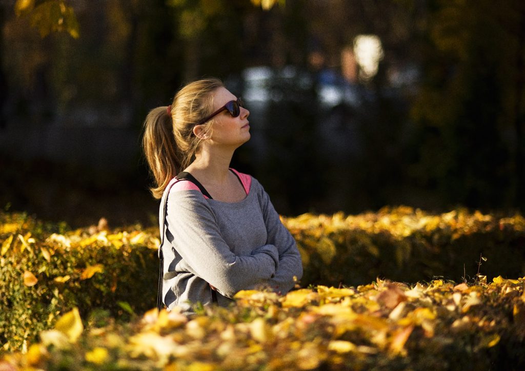 Portret kobiety w przeciwsłonecznych okularach. Kobieta stoi pomiędzy żywopłotem pokrytym jesiennymi liśćmi.