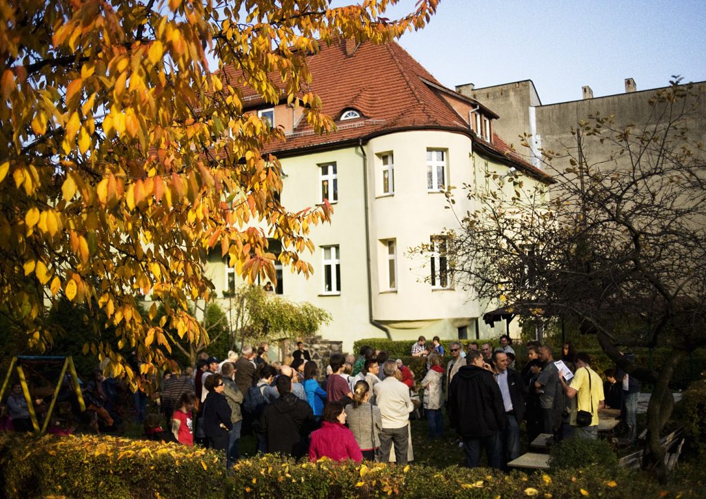Grupa os&oacute;b zgromadzona w ogrodzie przed jasną kamienicą z czerwonym dachem. Na pierwszym planie jesienne drzewo o ż&oacute;łtych liściach i niski żywopłot.