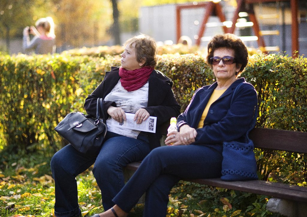 Dwie kobiety siedzą na ławce w parku. Jedna z nich trzyma w ręku gazetę, druga w słonecznych okularach patrzy w obiektyw. W tle jesienny park.