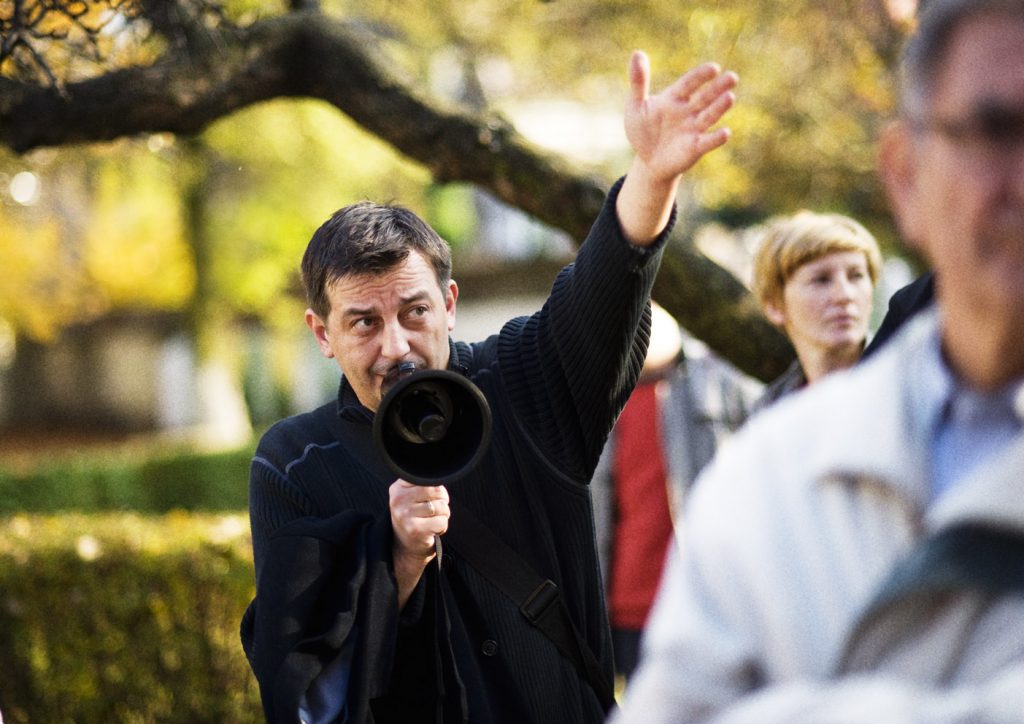 Mężczyzna m&oacute;wi przez megafon i wskazuje kierunek ręką. W tle rozmyte postacie ludzi i drzew w jesiennym parku.