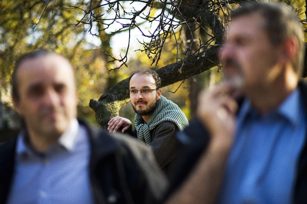 Mężczyzna w okularach i narzuconym na ramiona swetrze w paski, oparty o drzewo w jesiennym parku, ujęty pomiędzy dwiema nieostrymi postaciami na pierwszym planie.