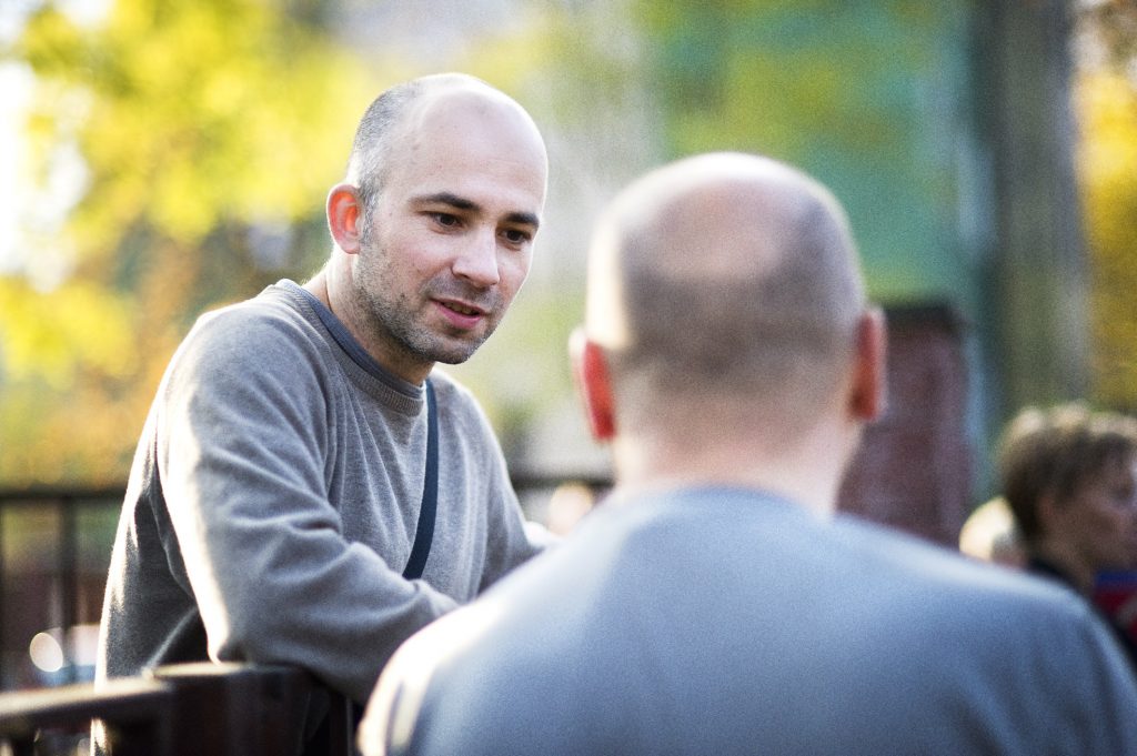 Mężczyzna z kr&oacute;tkim zarostem w szarym swetrze rozmawia z inną osobą, widoczną od tyłu, na tle jesiennego parku.