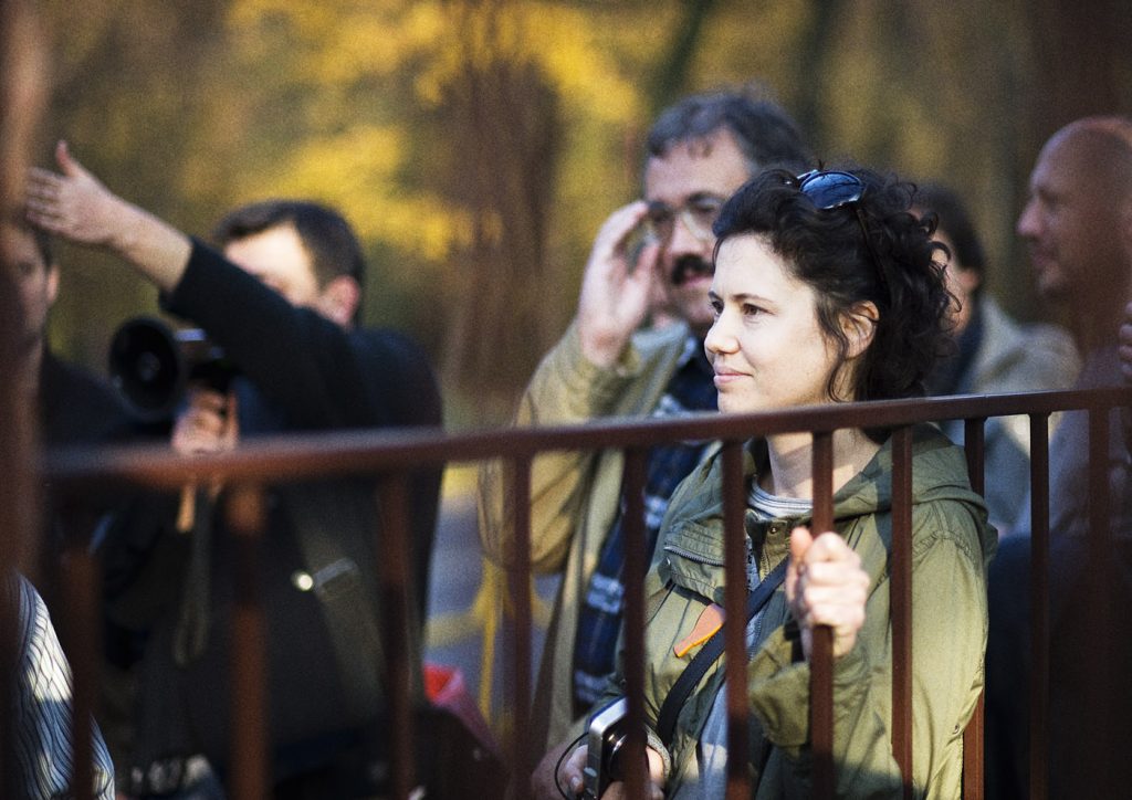 Młoda kobieta z profilu trzyma jedną rękę na barierce. W tle kilku mężczyzn, jeden z nich m&oacute;wi przez megafon.