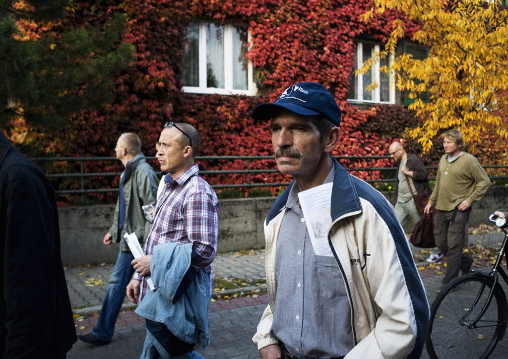 Kilku mężczyzn idzie chodnikiem. W tle ściana z winoroślą w czerwonym kolorze.