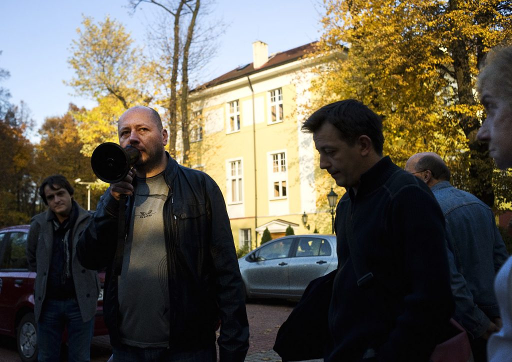 Mężczyzna w sk&oacute;rzanej kurtce przemawia m&oacute;wi do megafonu. W koło niego kilka os&oacute;b. W tle ż&oacute;łty budynek i jesienne drzewa.