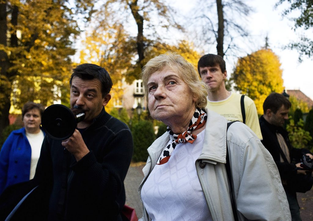 Starsza kobieta w jasnej kurtce i wzorzystej apaszce spogląda w g&oacute;rę, stoi obok mężczyzny m&oacute;wiącego przez megafon. W tle jesienny park.