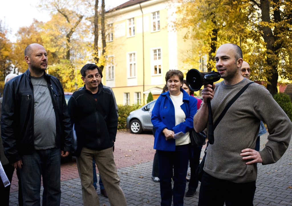 Mężczyzna z megafonem w ręku. Za nim kilka dorosłych os&oacute;b, na tle ż&oacute;łtego budynku i jesiennych drzew.