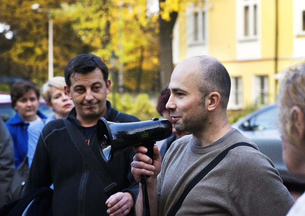 Mężczyzna trzyma w ręku czarny megafon. W koło niego stoi kilka dorosłych os&oacute;b. W tle jesienny park.