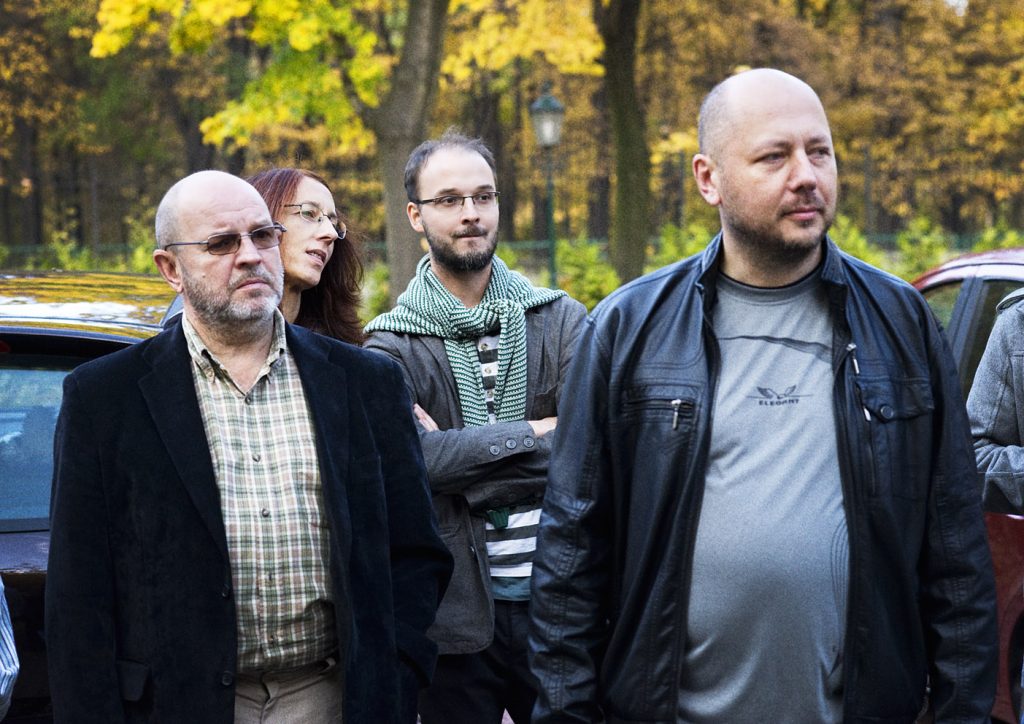 Czworo dorosłych os&oacute;b w jesiennym parku; na pierwszym planie mężczyzna w czarnej sk&oacute;rzanej kurtce oraz starszy mężczyzna w ciemnej marynarce i koszuli w kratę.