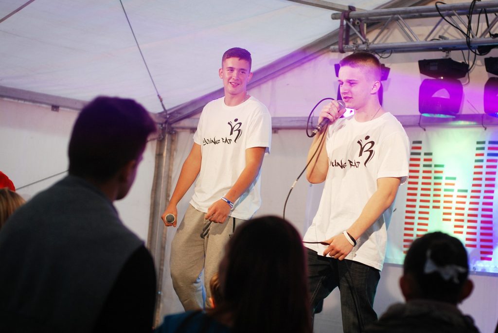 Dw&oacute;ch młodych wykonawc&oacute;w w białych koszulkach na scenie pod namiotem. Na pierwszym planie plecy zgromadzonej publiczności. Kolejne ujęcie.