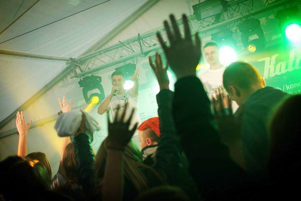 Uniesione ręce publiczności. W tle dwaj artyści na scenie w świetle reflektor&oacute;w.