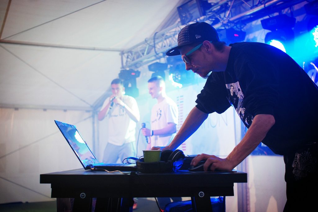 Mężczyzna w czapce i okularach przeciwsłonecznych pochyla się nad laptopem. W tle dwaj młodzi artyści w oświetleniu kolorowych reflektor&oacute;w.