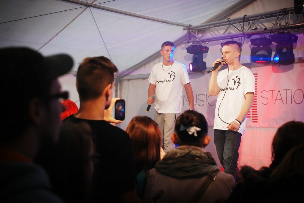 Dw&oacute;ch młodych wykonawc&oacute;w w białych koszulkach na scenie pod namiotem. Na pierwszym planie plecy zgromadzonej publiczności.