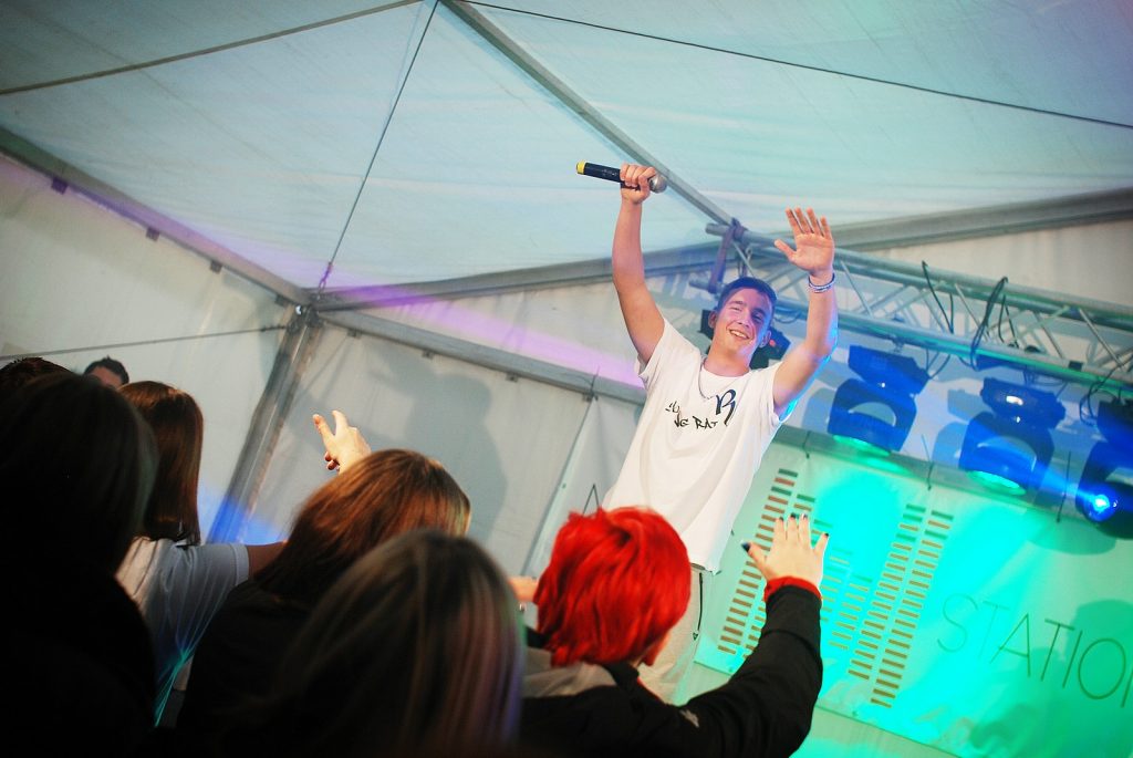 Młody wykonawca w białej koszulce występuje na scenie pod namiotem. W tle widoczny fragment napisu STATION oraz kolorowe oświetlenie sceniczne, a na pierwszym planie plecy zgromadzonej publiczności.