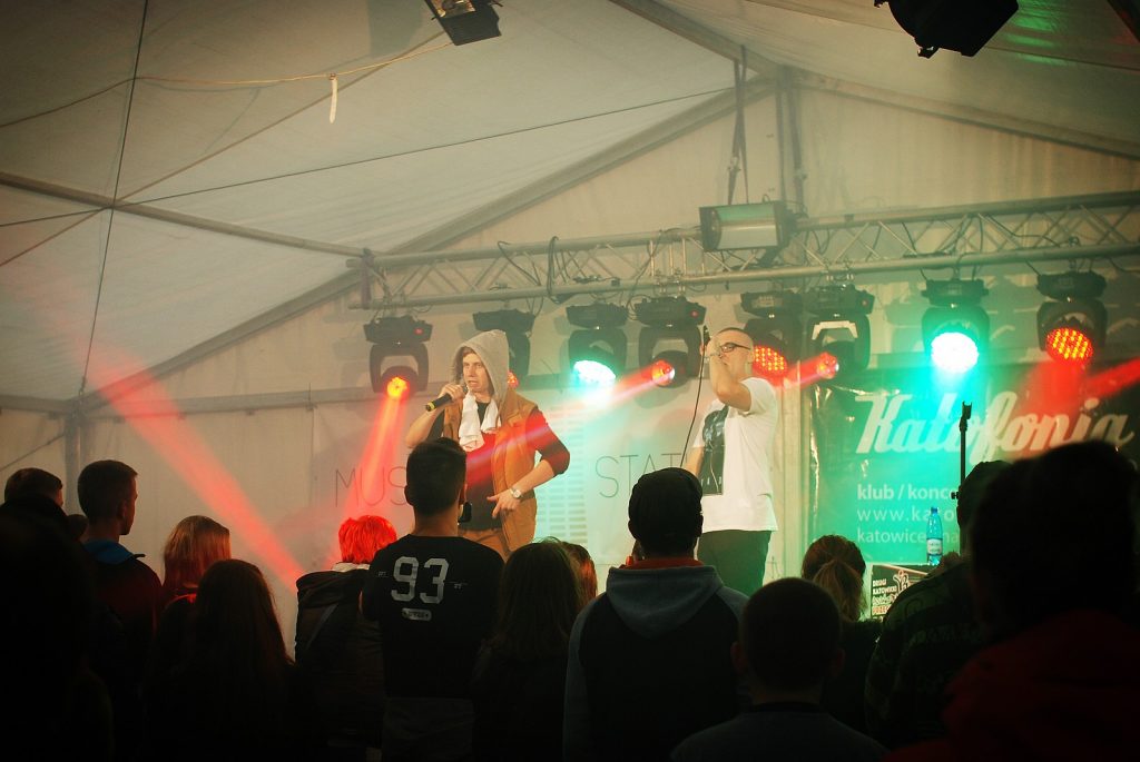 Widok zza plec&oacute;w publiczności na dw&oacute;ch artyst&oacute;w na scenie. Mężczyźni są oświetleni czerwonym światłem reflektor&oacute;w.