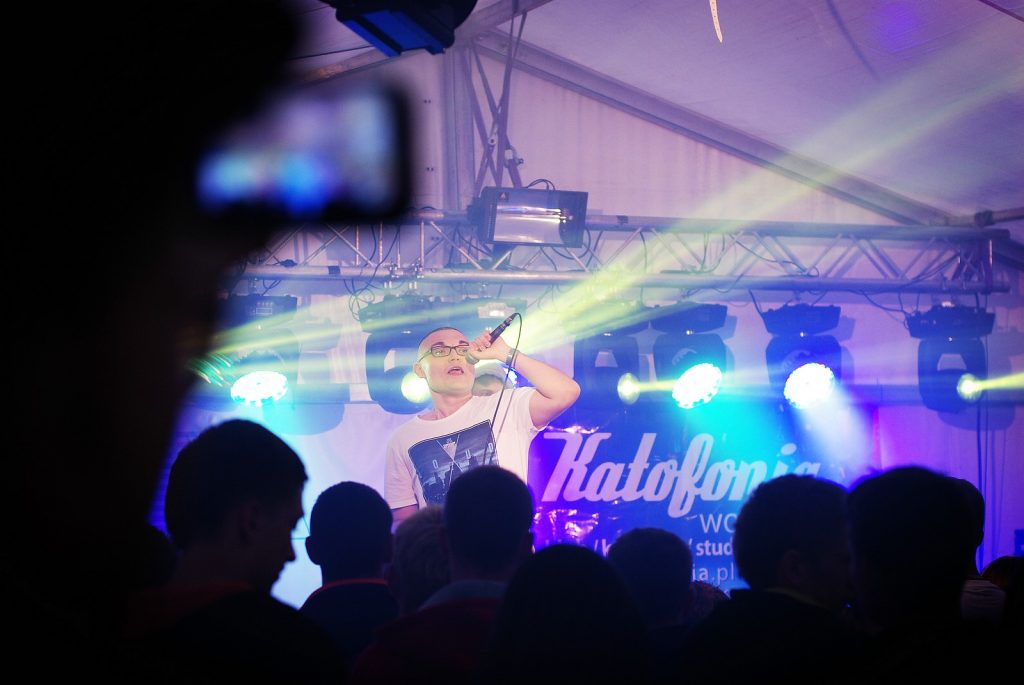Widok zza plec&oacute;w publiczności na mężczyznę na scenie. W tle kolorowe reflektory i baner z napisem Katofonia.