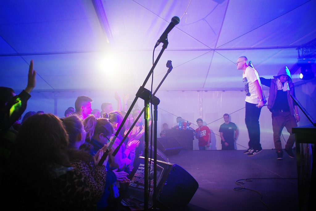 Na gł&oacute;wnym planie dwa mikrofony estradowe, w tle dwie osoby na scenie i publiczność.