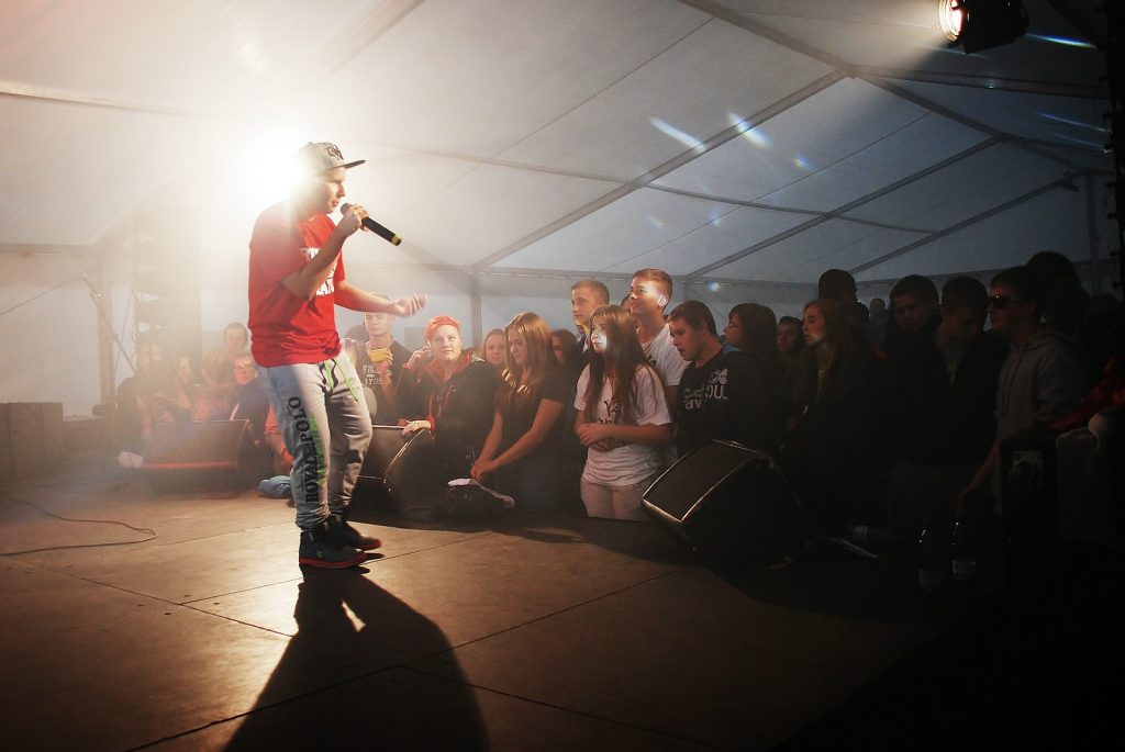 Mężczyzna w czerwonej koszulce, czapce na głowie stoi na estradzie. Przed nim bawi się publiczność.