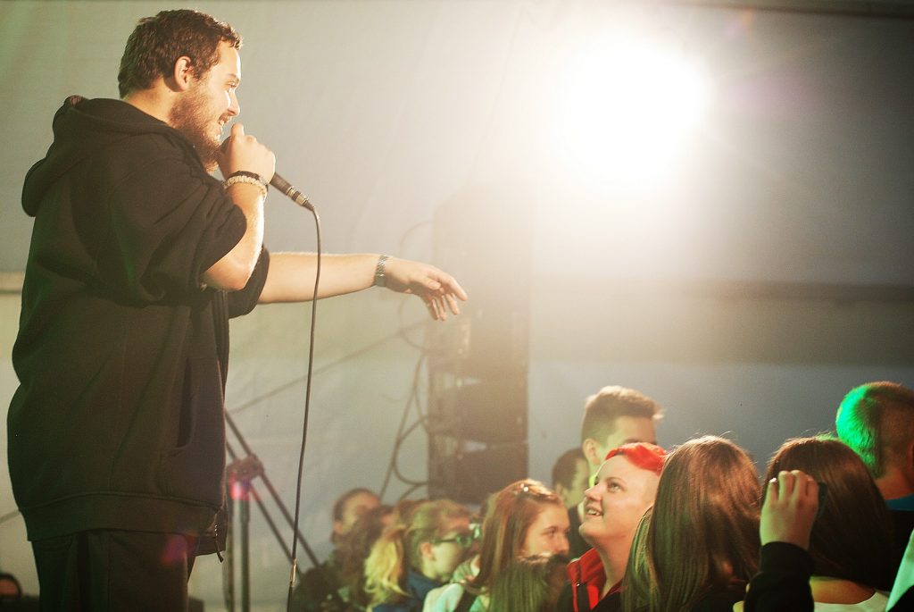 Na pierwszym planie mężczyzna w czarnej bluzie i mikrofonem w ręku stoi przed publicznością.