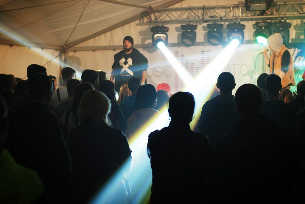 Koncert w namiocie festiwalowym. Widok zza plec&oacute;w publiczności na oświetloną reflektorami scenę, na kt&oacute;rej występuje dw&oacute;ch artyst&oacute;w. Jeden z nich ma na czarnej bluzie biały symbol skrzyżowanych młotk&oacute;w g&oacute;rniczych.