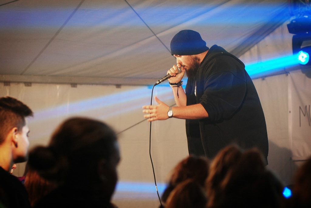 Mężczyzna w czarnej bluzie i czarnej czapce z mikrofonem w ręku. W tle niebieskie światło estradowe.