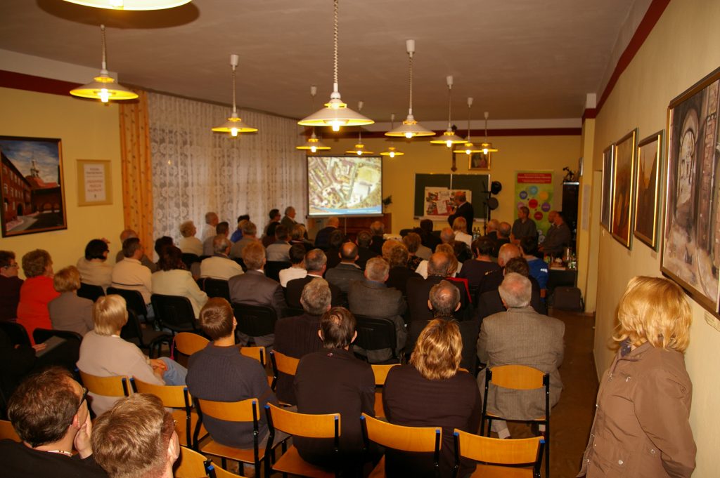 Widok od tyłu na salę wypełnioną ludźmi. Osoby siedzą na krzesłach i słuchają wypowiedzi prelegenta.