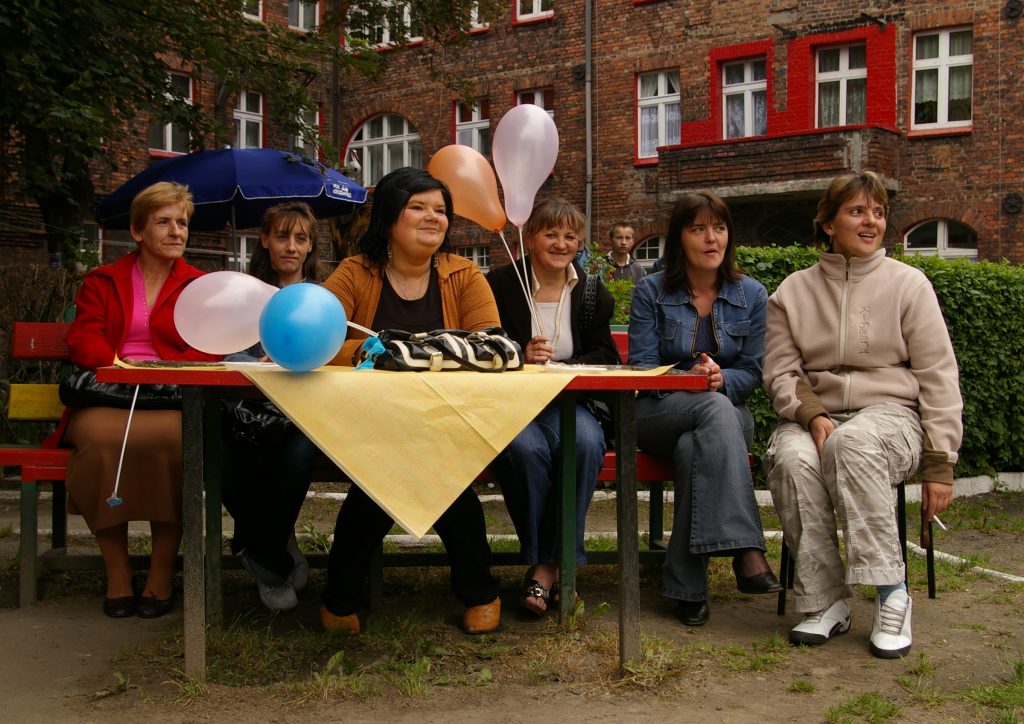 Grupa sześciu kobiet siedzi za stołem ustawionym na podw&oacute;rku przed ceglanym budynkiem. Kilka z nich trzyma kolorowe balony.