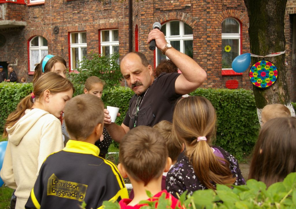 Mężczyzna unosi rękę do g&oacute;ry w kt&oacute;rej trzyma mikrofon. W koło niego grupa dzieci. W tle ceglany budynek.