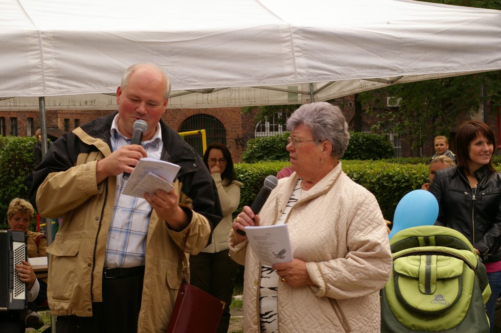 Kobieta i mężczyzna z mikrofonami w rękach stoją przed białym namiotem. W tle kilka os&oacute;b i ceglany budynek.