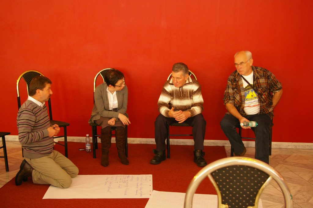 Grupa czterech os&oacute;b podczas warsztat&oacute;w na tle czerwonej ściany. Jeden mężczyzna klęczy przy arkuszu papieru na podłodze, a trzy osoby siedzą na krzesłach i obserwują go.