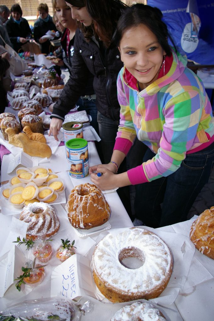 Młoda kobieta kolorowej bluzie w kratę przy stole zastawionym domowymi wypiekami. W tle kilka os&oacute;b przy stoisku.
