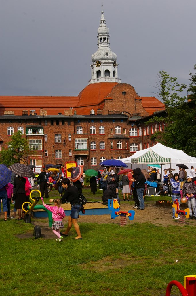 Grupa ludzi z parasolami na świeżym powietrzu. W tle kamienica Nikiszowca i kości&oacute;ł Św. Anny.