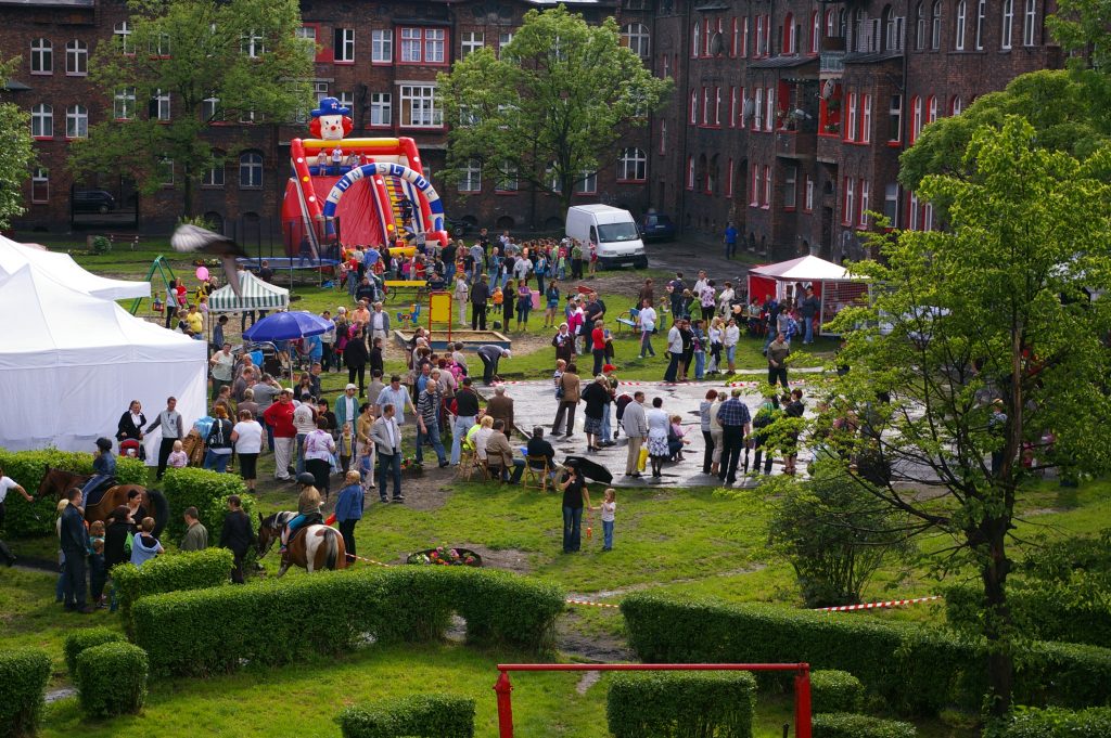 Widok z g&oacute;ry na wydarzenie plenerowe między ceglanymi kamienicami. W tle duża, dmuchana zjeżdżalnia z głową klauna, wok&oacute;ł kt&oacute;rej gromadzą się dorośli i dzieci.