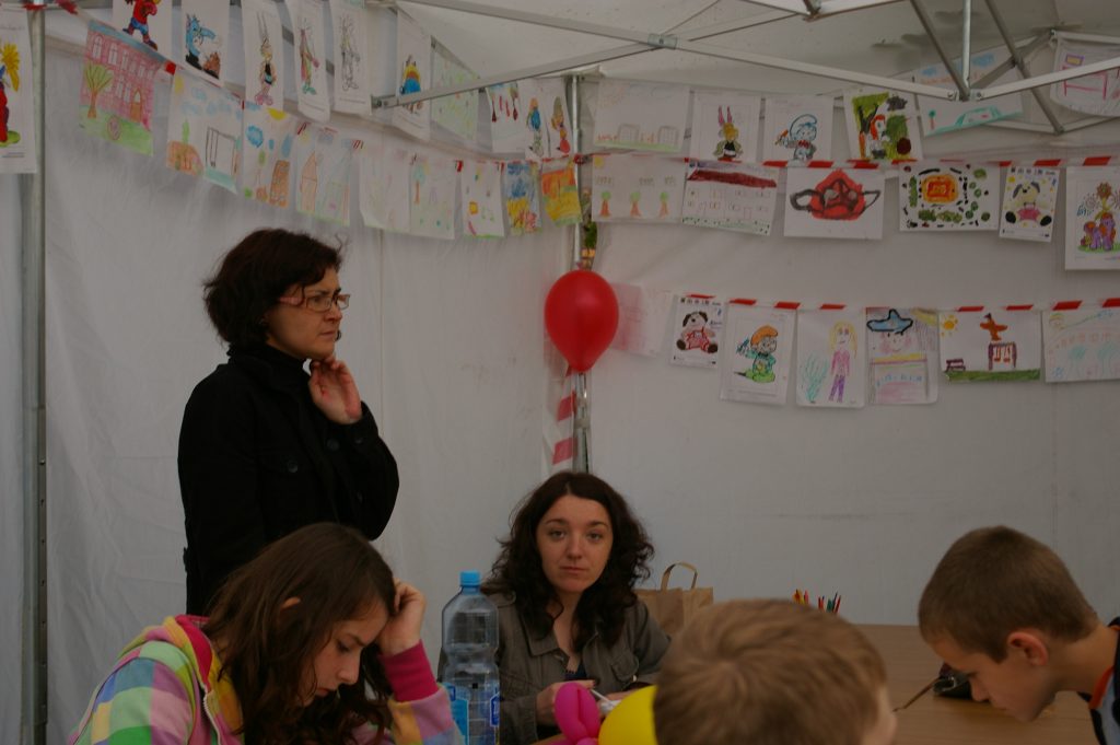 Wnętrze namiotu udekorowane dziecięcymi rysunkami i czerwonym balonem. Dwie kobiety obserwują dzieci siedzące przy stole i zajęte pracą plastyczną.