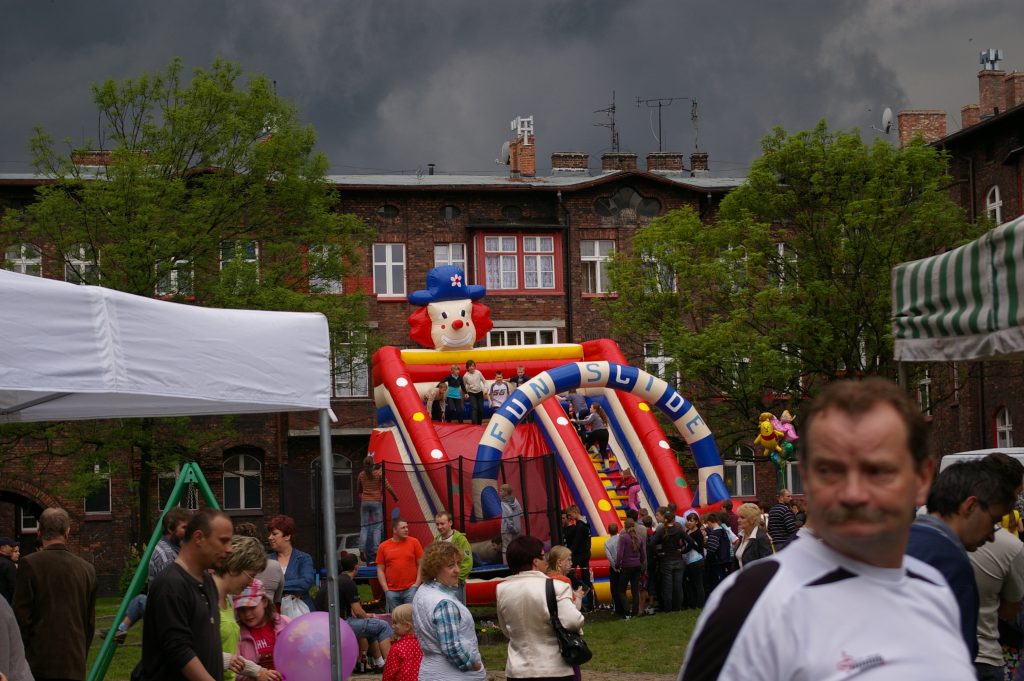 Festyn osiedlowy na tle ceglanych kamienic pod zachmurzonym niebem. Dzieci bawią się na dmuchanej zjeżdżalni w kształcie klauna otoczonej przez tłum ludzi.