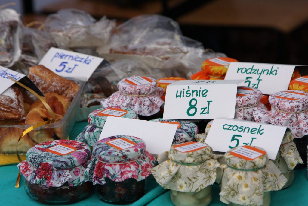 Stoisko z pierniczkami, domowymi przetworami w słoiczkach udekorowanych wzorzystymi materiałami na nakrętkach. Na kartkach zapisane ceny produkt&oacute;w.