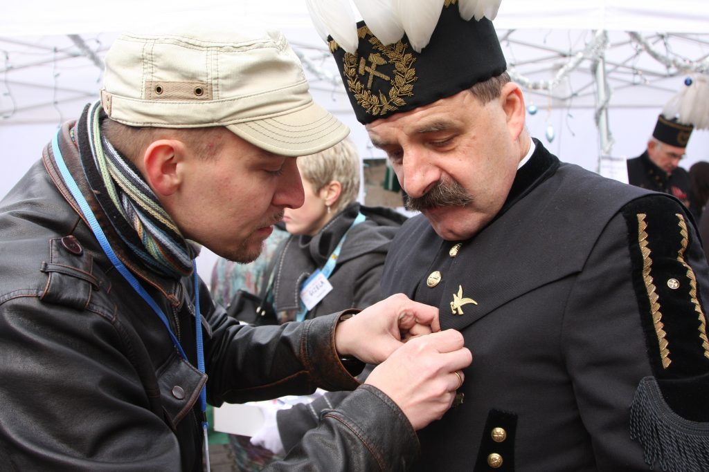 Mężczyzna przypina odznakę drugiemu mężczyźnie, kt&oacute;ry jest ubrany w str&oacute;j galowy g&oacute;rnika. W tle widać inne osoby i zadaszenie namiot&oacute;w.
