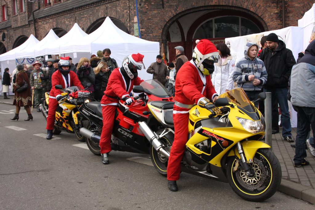 Trzech motocyklist&oacute;w przebranych w stroje mikołaja. W tle uczestnicy jarmarku i białe namioty wystawowe.