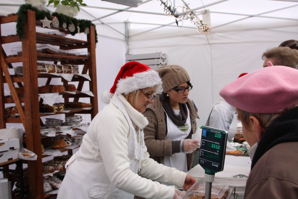Dwie kobiety sprzedają świąteczne wypieki, rozmawiają z kupującymi. W tle regał z ciastami.