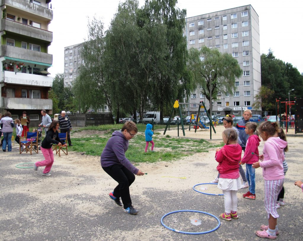 Grupa dzieci w trakcie zabawy zręcznościowej na świeżym powietrzu. W tle dorosłe osoby, miejska zieleń i bloki mieszkalne.