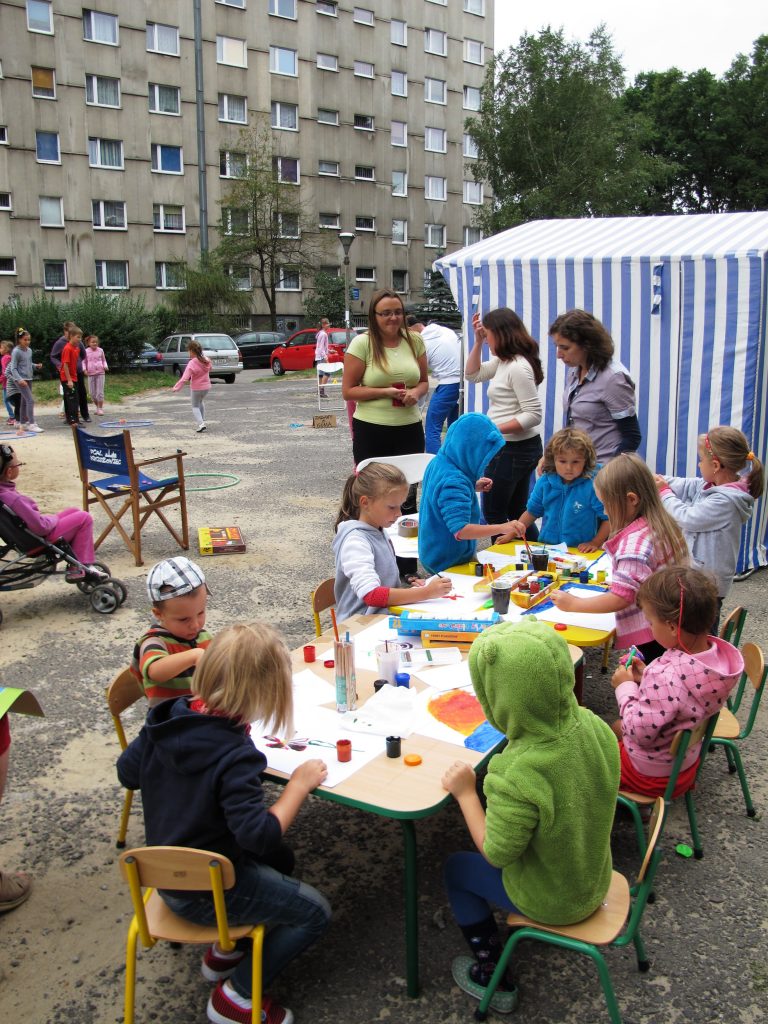 Grupa dzieci, siedzi przy stolikach rozstawionych na świeżym powietrzu. W tle namiot ogrodowy, rozmawiający dorośli, bawiące się dzieci na placu przed blokiem.