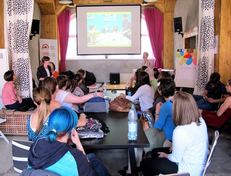 Grupa os&oacute;b siedzi przy stole z głowami skierowanymi na ekran z prezentacją.