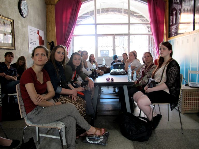 Grupa młodych os&oacute;b siedzi przy długim stole w jasnej sali z dużym oknem i r&oacute;żowymi zasłonami.