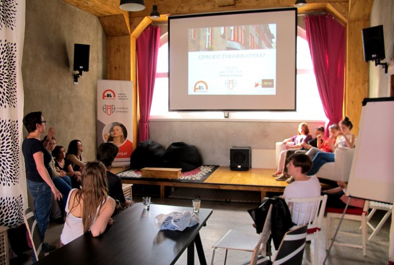 Grupa os&oacute;b w sali z ekranem na kt&oacute;rym wyświetlony jest napis Czym jest żywa biblioteka. W tle widać baner Centrum Zimbardo.