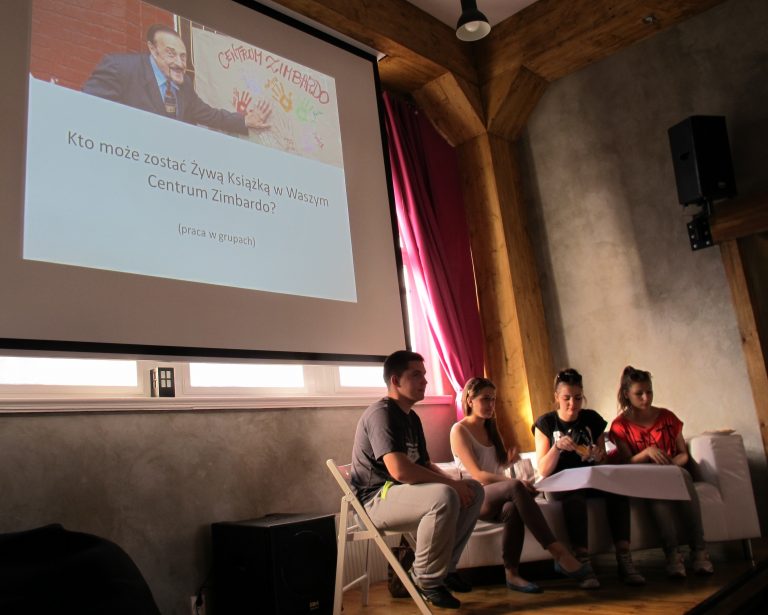 Grupa młodych ludzi siedzi w sali przed ekranem z prezentacją pt. &bdquo;Kto może zostać Żywą Książką w Waszym Centrum Zimbardo?&rdquo;