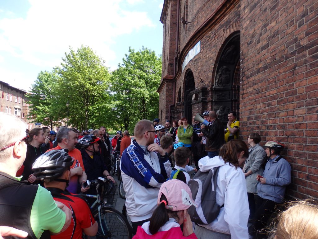 Grupa rowerzyst&oacute;w słuchająca przem&oacute;wienia przez megafon przed wejściem do zabytkowego, ceglanego budynku w Nikiszowcu.