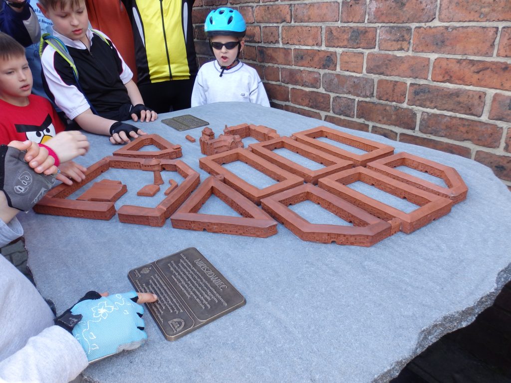 Tr&oacute;jka dzieci stoi przy makiecie dotykowej osiedla Nikiszowiec. Na pierwszym planie dłoń dotyka tabliczki informacyjnej z opisem.