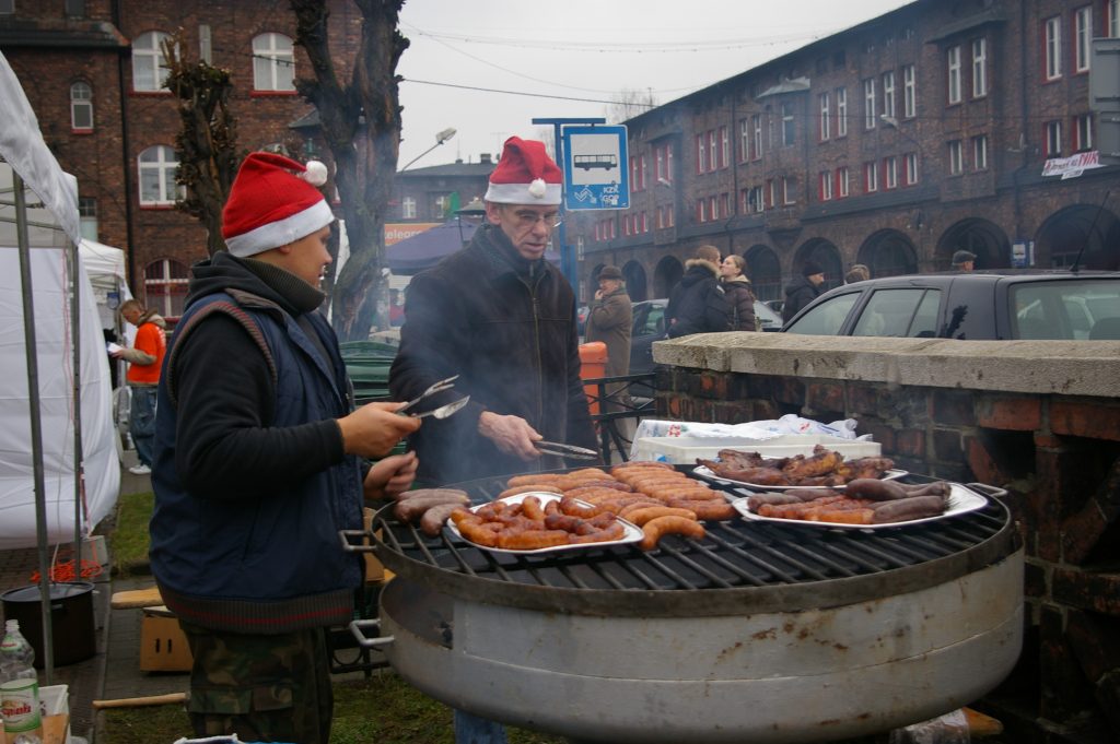 Mężczyzna i chłopiec stoją przy grillu z kiełbaskami. W tle całująca się para.