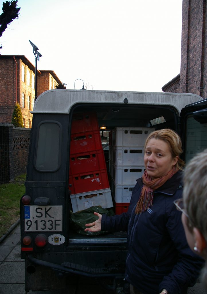 Dwie kobiety rozmawiają, w tle bagażnik samochodu załadowany skrzynkami.